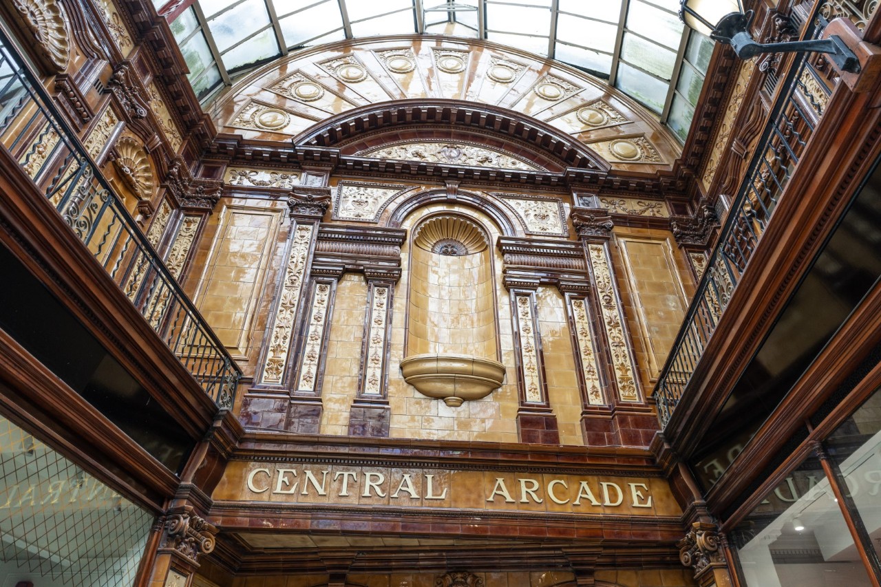 A wooden-panelled courtyard of a two-story shopping arcade with a semicircular glass roof. The word “Central Arcade” is written in gold letters on a centrally placed wooden panel. The upper floor is equipped with railings.