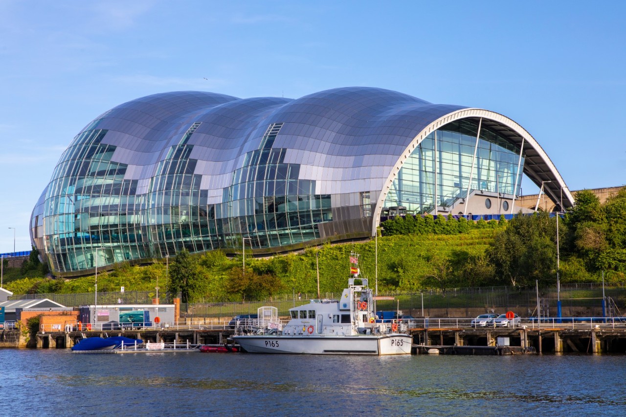 Futuristic, wave-like building with a completely glass exterior façade. It stands on a bank, with several smaller boats moored on the shore.