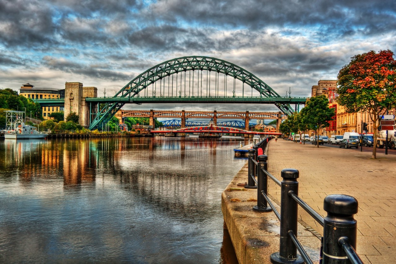 A large, green steel arch bridge spans a river; in the background, another red steel bridge can be seen. To the left of the bridge, a historic building is visible. To the right, a paved path leads along the bank; a fence with pillars forms the border to the water's edge; an autumn-coloured tree stands on the riverside path; behind it, several houses are visible.