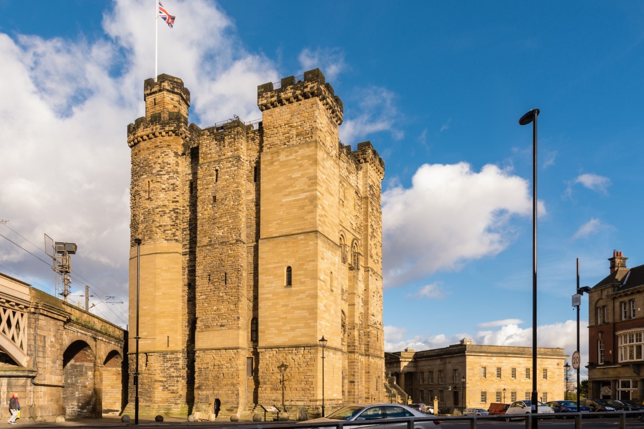 A massive, sandstone-coloured, angular castle with towers in the corners, which are crenellated. There is a flag on the left tower. To the left of the castle is an old bridge with archways, and to the right of the castle are further buildings.
