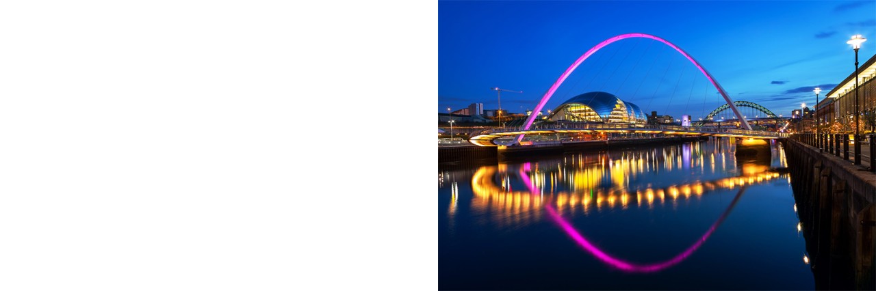 Evening shot of a river in a city, with a pink-lit arched bridge in the foreground, reflected in the water. More lights from the adjacent buildings are reflected in the water. A modern building is visible in the bridge arch, with an additional arched bridge in the background. On the right, in the foreground, there is another illuminated building right on the edge of the riverbank.