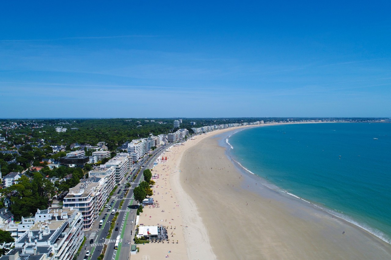 The picture shows a long, curved sandy beach, with a promenade of white houses on the left, between which numerous green trees protrude. The water on the right is turquoise blue and the sky is cloudless. 