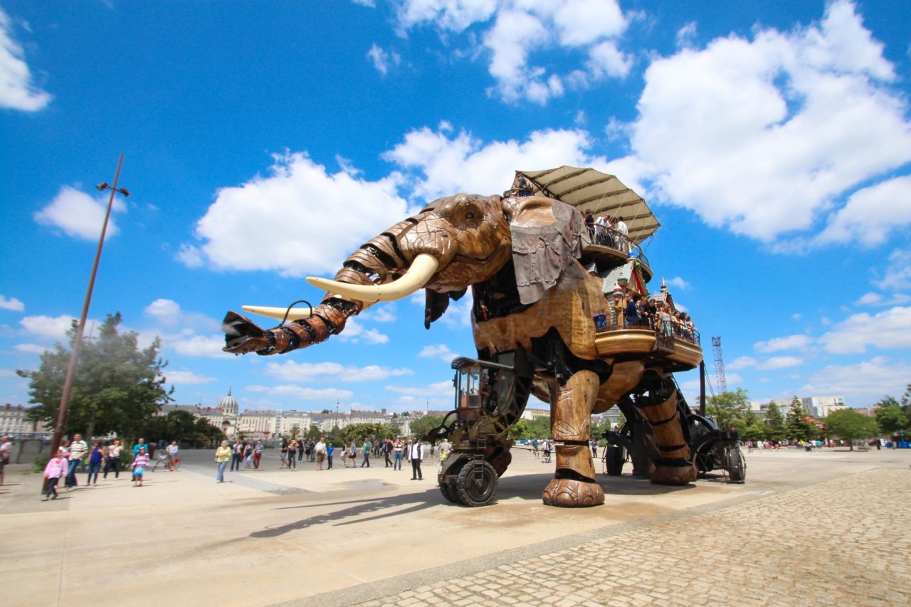 The picture shows a square with a huge elephant made of wood and metal, with people standing at its feet. On the elephant's back is a platform on which people are standing. 