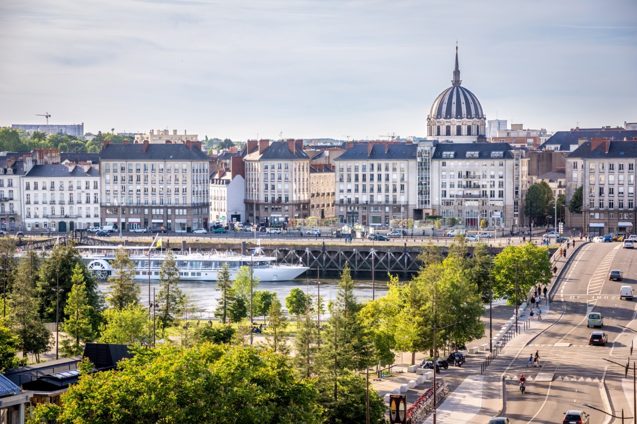 The picture shows a city view by the river. Trees and a multi-lane road can be seen in the foreground. In the background are buildings with classical facades and a striking domed building. An excursion boat is moored on the riverbank.