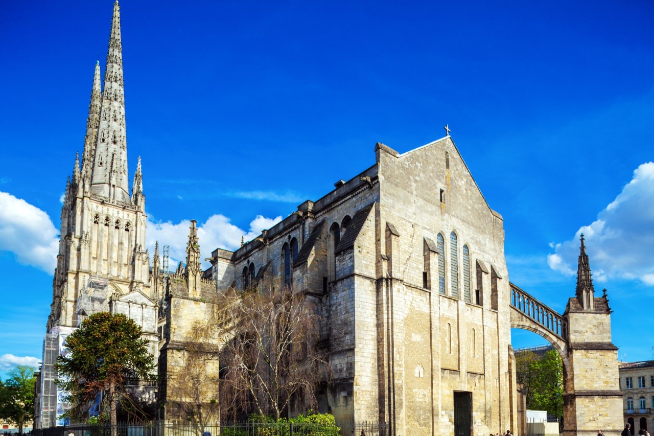 Side view of the exterior of a Gothic cathedral. In the centre of the picture is the nave built of light, sand-coloured stone with arched windows, which is connected to a small tower on the right by a slightly sloping bridge. On the left there are two pointed main towers of the cathedral, which are decorated with intricate stone carvings. A bright blue sky with a few clouds can be seen in the background.