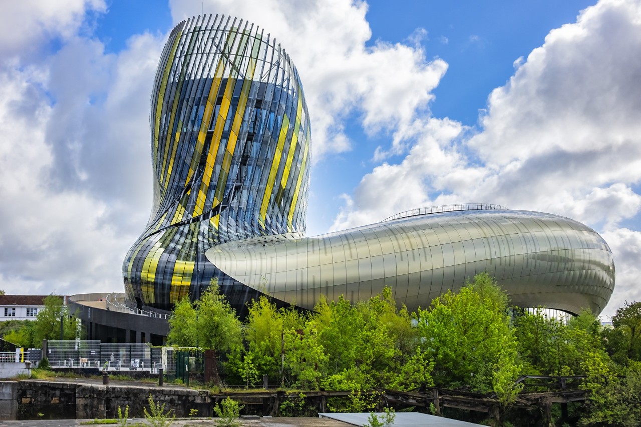 View of the curved glass and steel construction of a modern, futuristic-looking building. The curved shape is reminiscent of a wine decanter. The glass windows in the part of the building that rises upwards shimmer blue, yellow and green. The tube-like part of the building leading to the right is encased in steel. There are trees in front of the building. Clouds and some blue sky can be seen in the background.