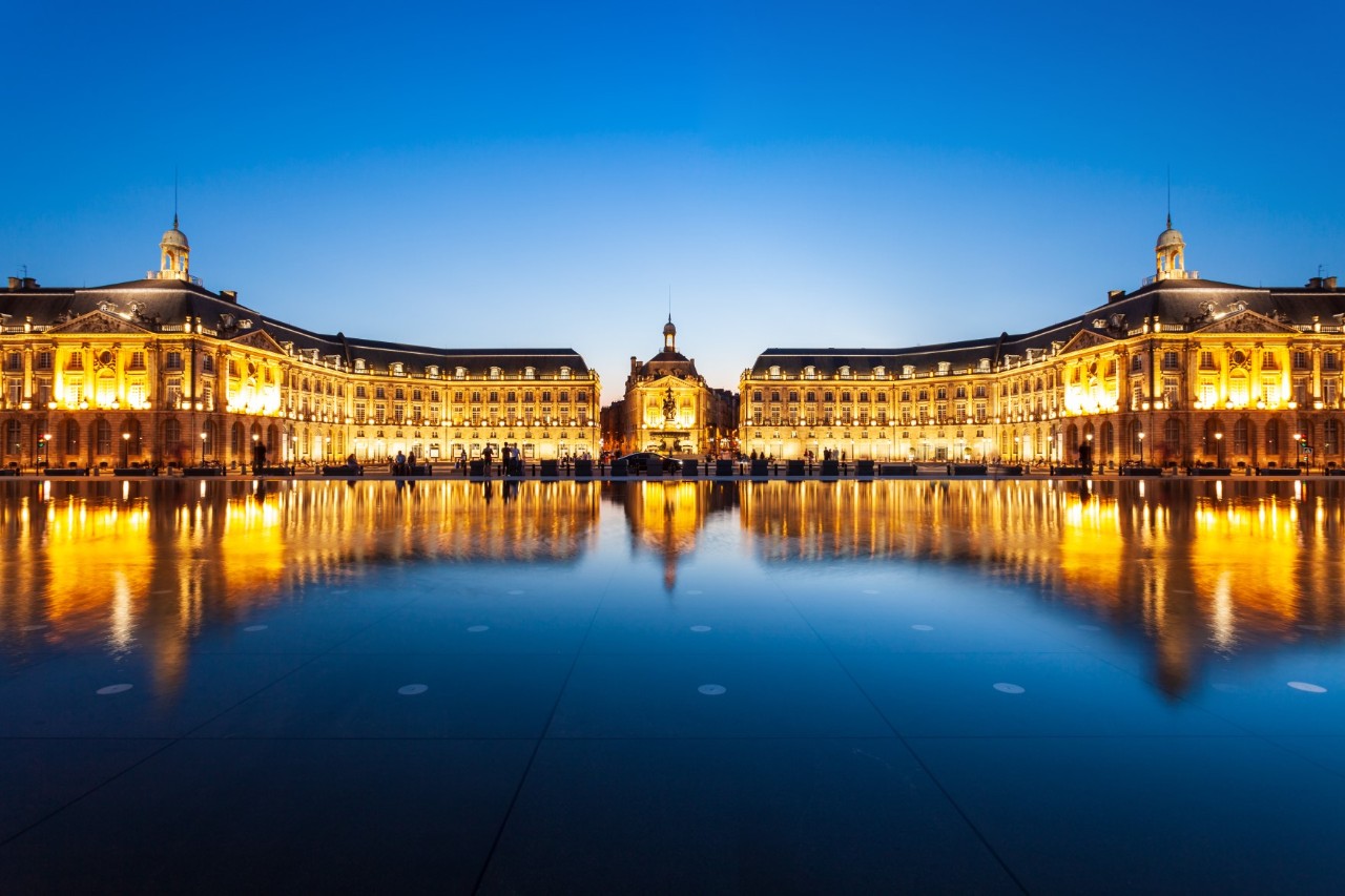 View of a magnificent, symmetrical, brightly lit palace building at dusk. In front of the building is a pool in which the buildings are reflected.