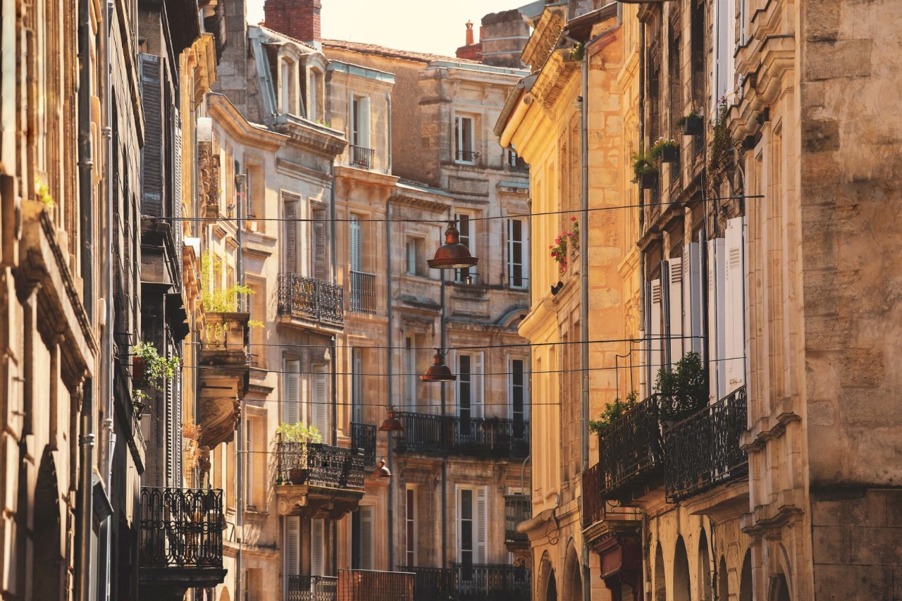 View of a colourful row of buildings in the old town, bathed in the warm, yellow light of the evening sun. Several planted balconies and bay windows can be seen on the buildings to the right and left of a narrow street. Washing lines stretched across the street are visible between the houses.