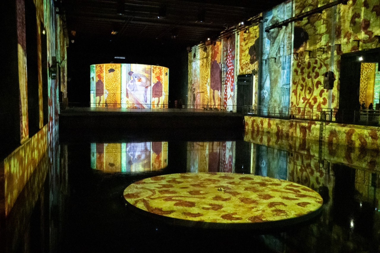 View into an underground bunker room with a pool in the centre. The room is dark and various works of art are projected onto the walls and the surface of the water, illuminating the room in bright colours.