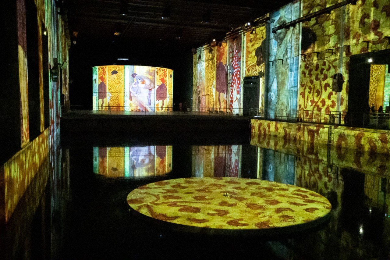 An underground bunker room with a pool in the centre. The room is dark and various works of art are projected onto the walls and the surface of the water, illuminating the room in bright colours.