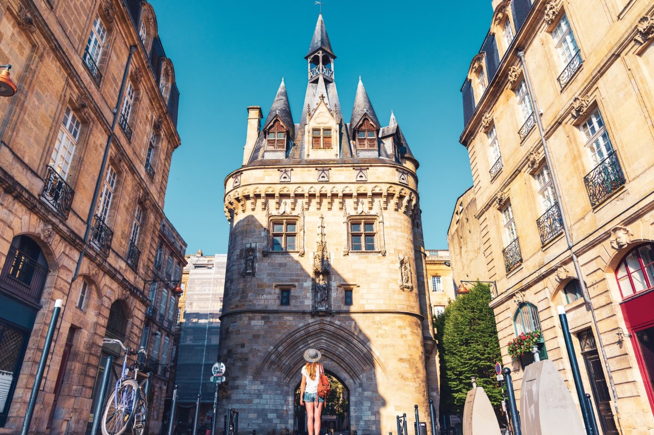 A large city gate lined with turrets, in front is a woman standing with her back to the picture, looking upwards. On both sides of the gate are historic, three-storey buildings with high windows and French balconies; a bicycle is parked on the left.