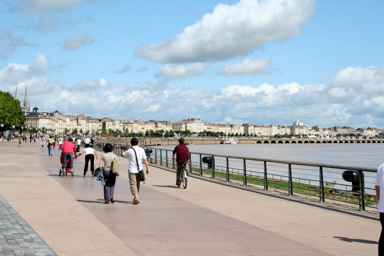 View of a wide waterside promenade where several people are walking and cycling along. A row of buildings and two church towers can be seen on the horizon and a river can be seen on the right, separated from the promenade by a fence.