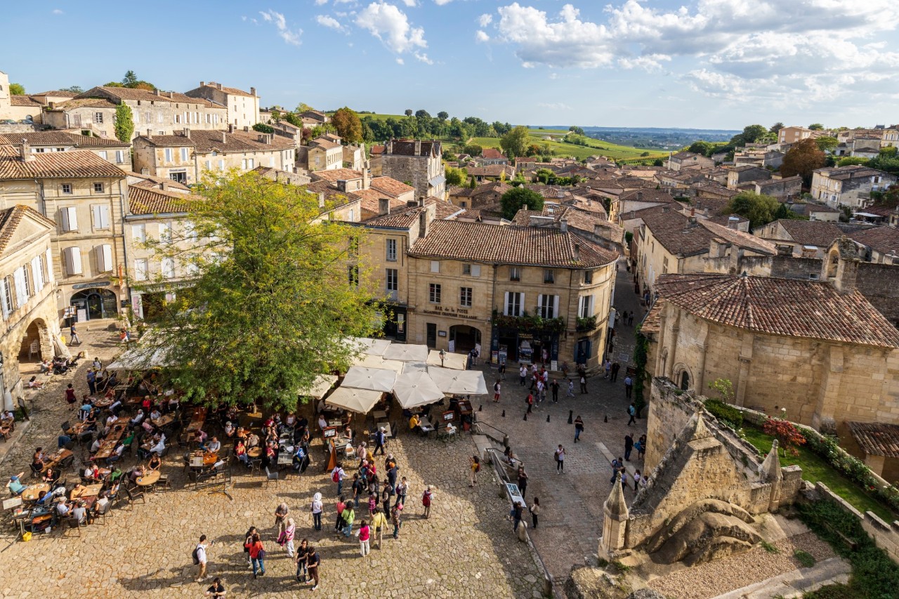 A central market square in a medieval-looking town with people, white-covered market stalls and a few tables with people sitting at them. The square is lined with historic buildings.
