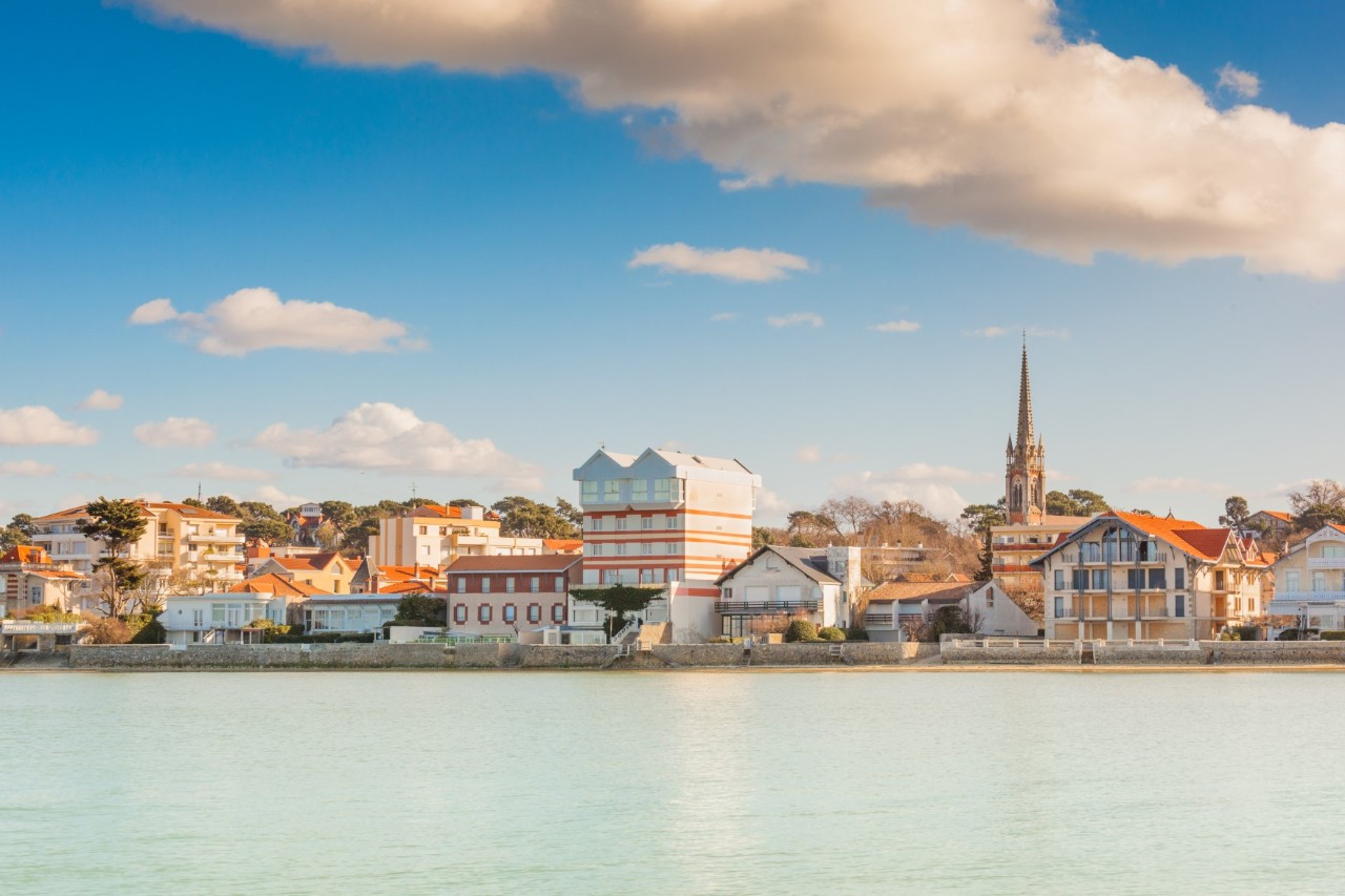 View from the waterside of a coastal town in the sunlight, directly bordering the sea. There are several buildings by the water and a church tower in the background. The blue sky is streaked with a few clouds, the light blue, almost pastel-coloured sea is in the foreground.