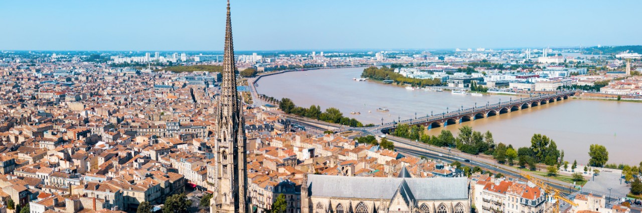 Aerial view of Bordeaux. In the centre is a Gothic cathedral with a long nave and a pointed tower in front of it, which are built on a large, empty square. There are numerous buildings with red roofs all around. A bridge crossing a river can be seen in the background. A blue strip of sky can be seen on the horizon.