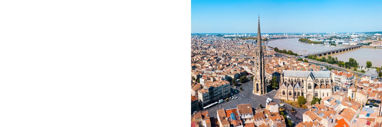 Aerial view of Bordeaux. In the centre is a Gothic cathedral with a long nave and a pointed tower in front of it, which are built on a large, empty square. There are numerous buildings with red roofs all around. A bridge crossing a river can be seen in the background. A blue strip of sky can be seen on the horizon.