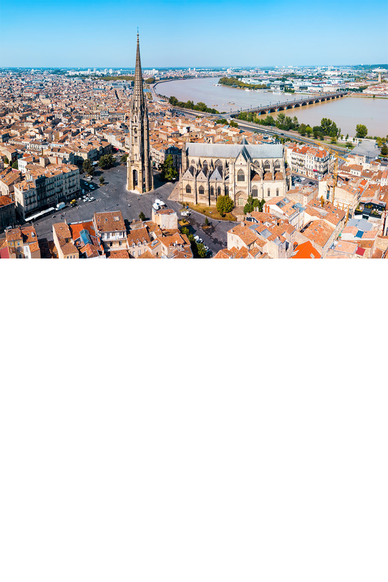Aerial view of Bordeaux. In the centre is a Gothic cathedral with a long nave and a pointed tower in front of it, which are built on a large, empty square. There are numerous buildings with red roofs all around. A bridge crossing a river can be seen in the background. A blue strip of sky can be seen on the horizon.