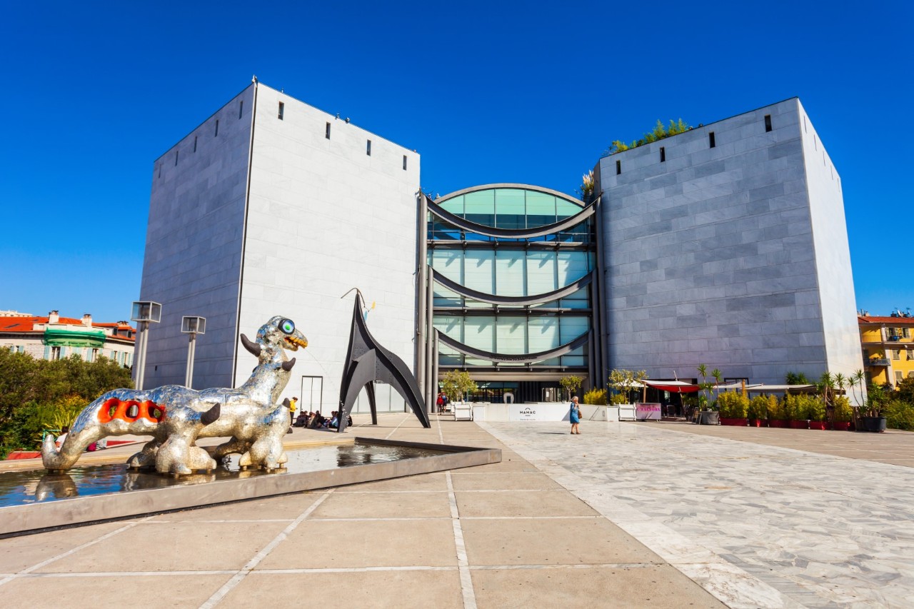 Exterior view of MAMAC, two large grey concrete blocks with glass entrance, fountain with sculpture in front, empty square 