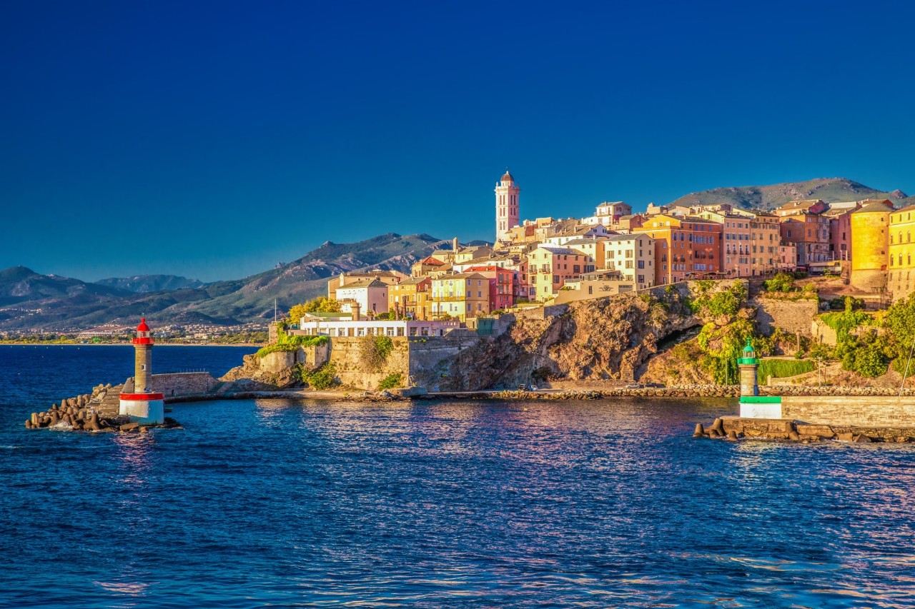 Buildings upon a rocky cliff. In front of it are two small lighthouses, each on a pier. A mountain panorama be seen in the background.