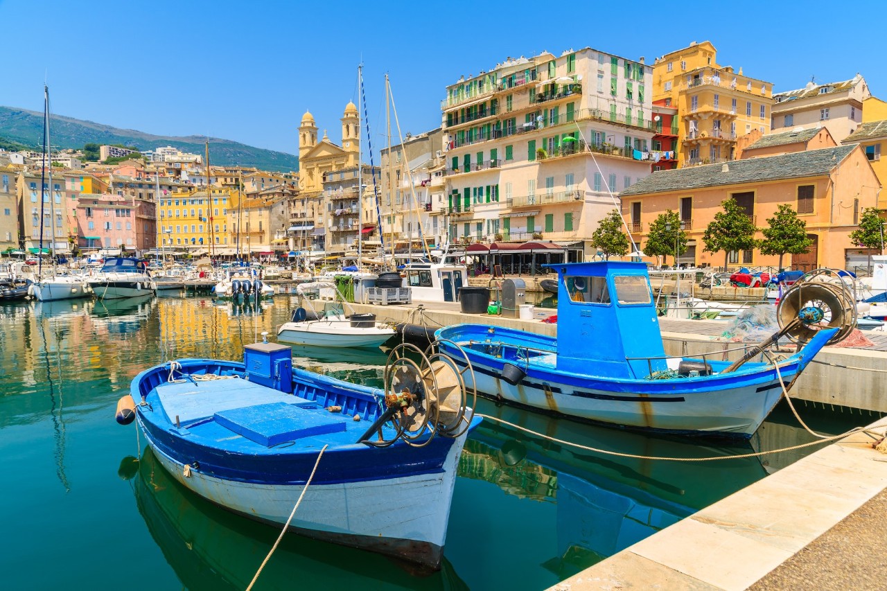 Harbour with two small, blue-white fishing boats in the foreground. Around the harbour basin are buildings with predominantly yellow fronts. The striking bell towers of a church can be seen a little further in the background.
