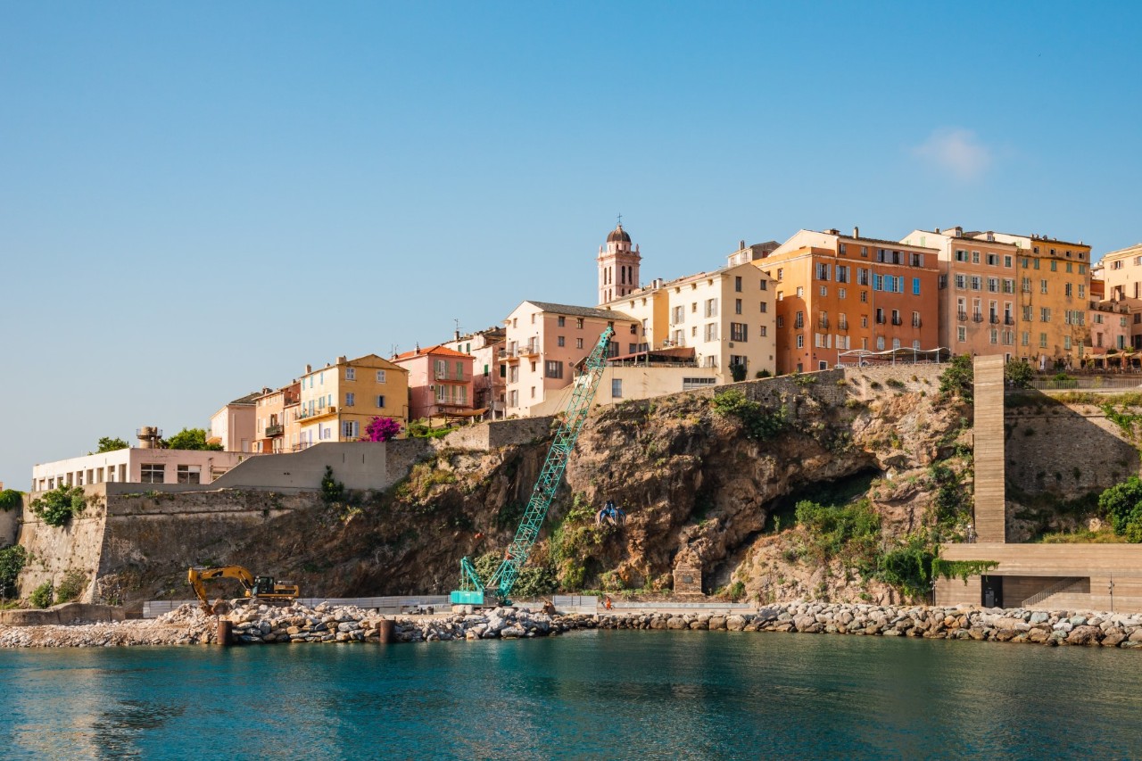 Sea of buildings upon a rocky cliff. A brown lift shaft can be seen on the right above a flat, rectangular building. Two construction vehicles are parked on the shore.