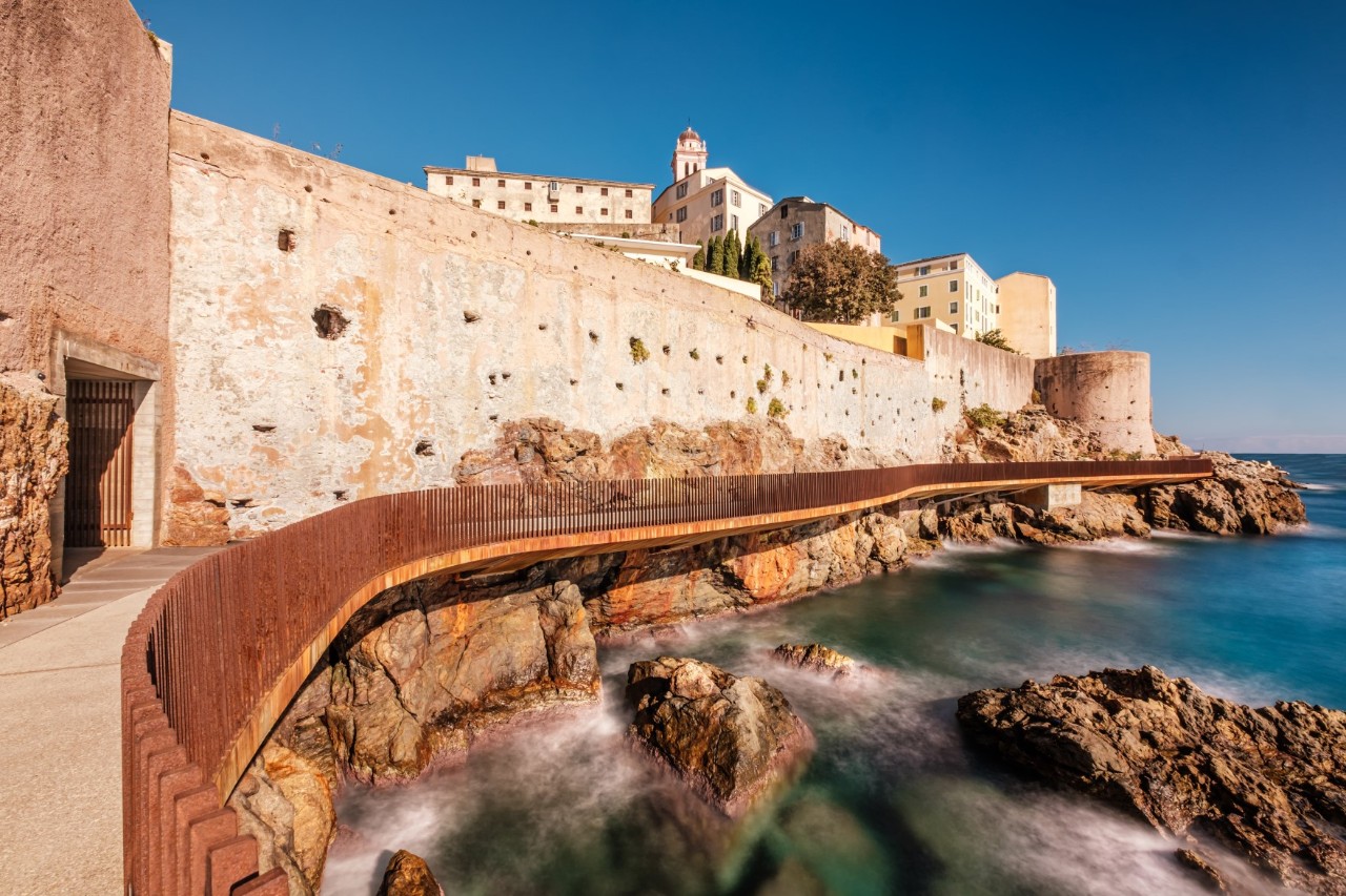 A deserted coastal path with a brown fence that stretches along a fortress wall directly above the sea. Stones in the water under the man-made path can be seen.