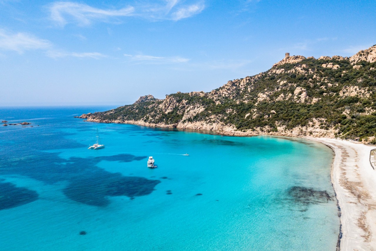 A bay with crystal-clear water on which two sailing boats are moored. Coral reefs can be seen on the sea floor. The light-coloured, narrow sandy beach on the right of the picture runs towards a green mountain.