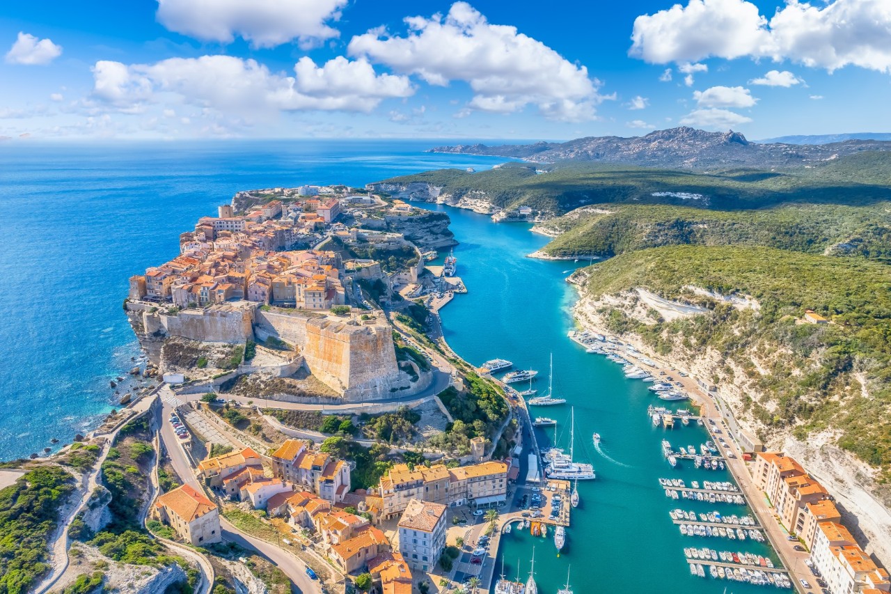 A town built on a rocky peninsula. To the right of the town, a harbour with many small boats can be seen bordering a built-up shoreline. The peninsula is surrounded by a green-blue sea and a green, hilly island side.