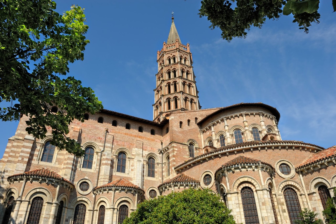 View from below of an impressive basilica made of red brick, with several arches and an angular bell tower. Green tree branches can also be seen.