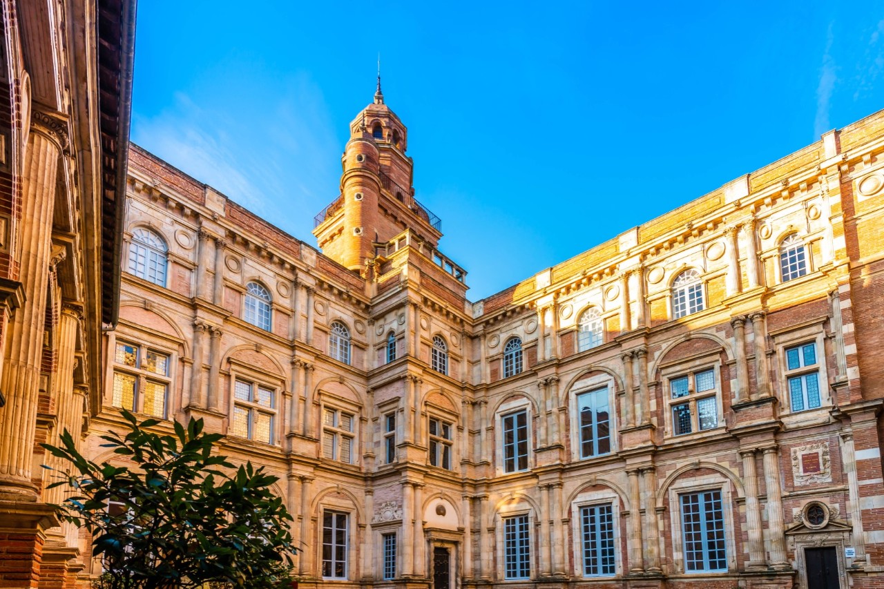 Corner view of a massive, three-storey building in a yellowish colour with large windows, embellishments on the front and a small tower on the roof. The sky is bright blue. There is a green bush in front of the building.