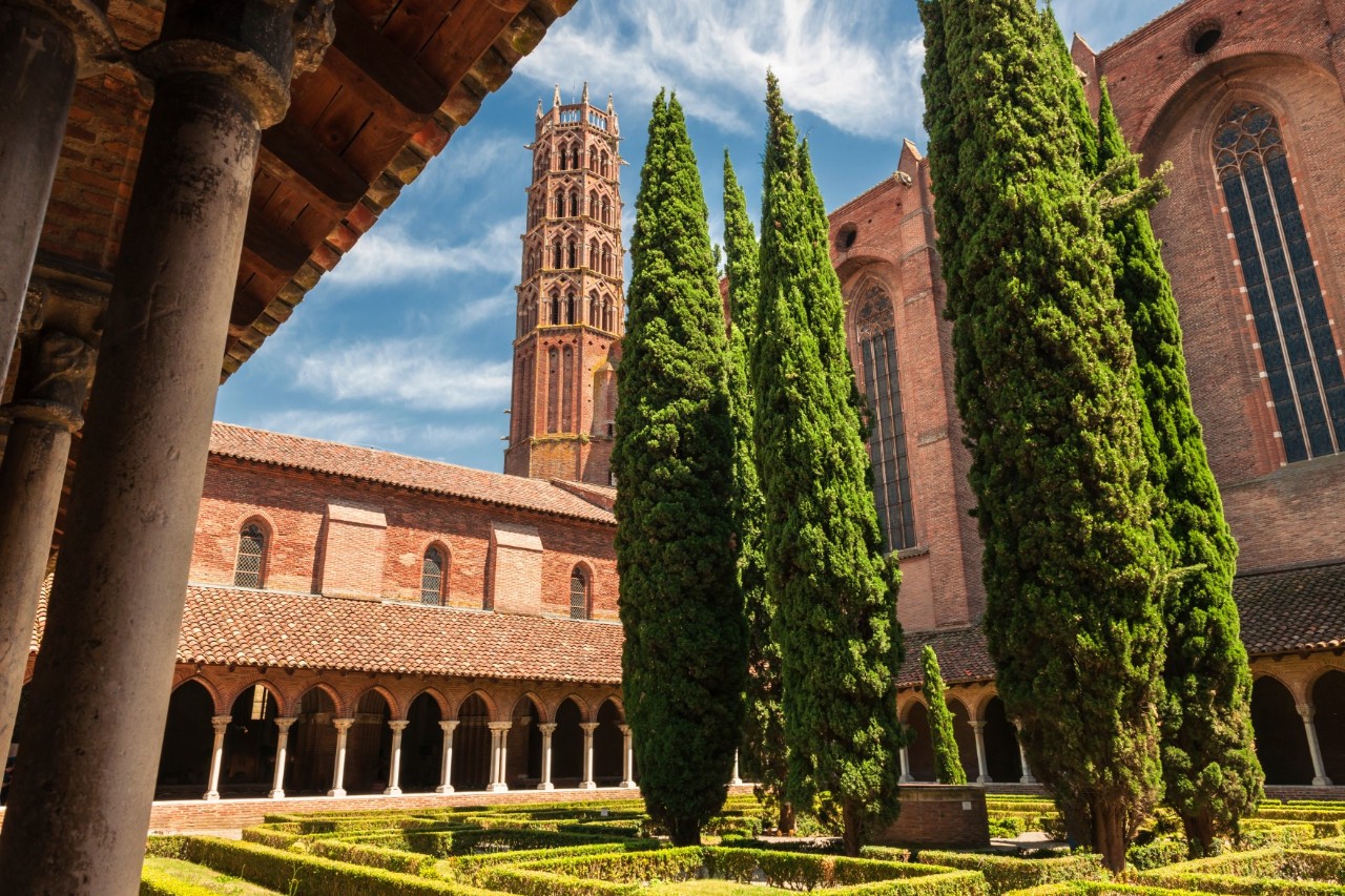 View from the inner courtyard of a striking red-brick church building with cloister and bell tower. The inner courtyard is landscaped with five narrow, tall, green trees and small hedges.