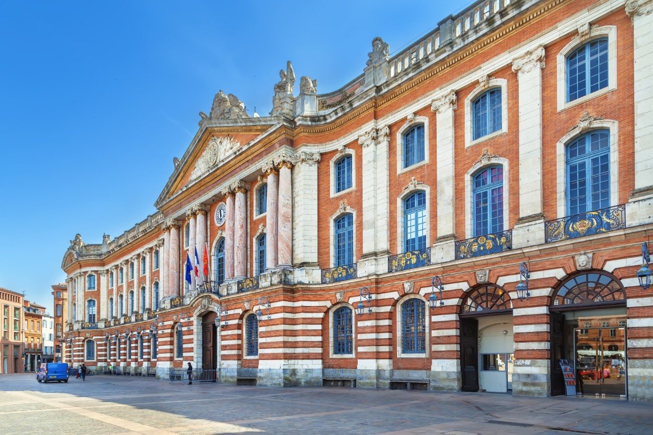 Front view of a striking red-and-white building with high windows. A blue vehicle is parked in front of the left end of the building.