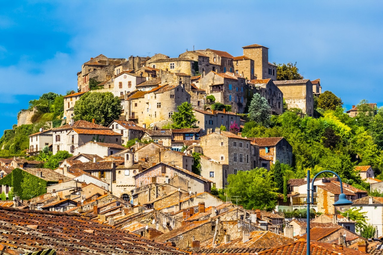 In the foreground is a view over red roofs to a densely built-up old village nestled against a mountain. There are green trees between the buildings.