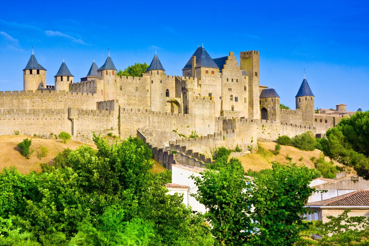 View of an imposing fortress with several watchtowers and a long wall. Trees and white buildings can be seen in the foreground.