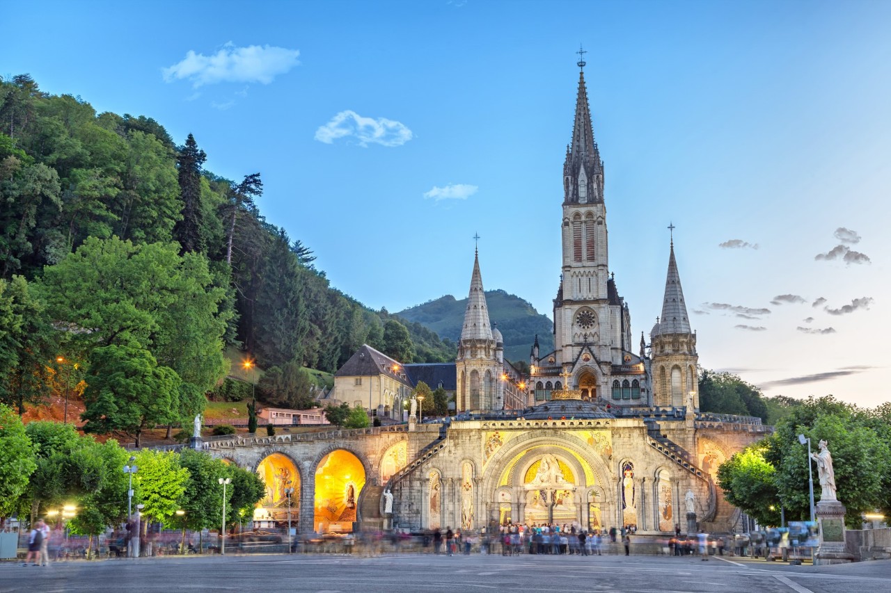 View of a basilica in the evening light, which is illuminated in the lower area and has pointed towers. The basilica is surrounded by trees and mountains. People can be seen blurred in front of the entrance.