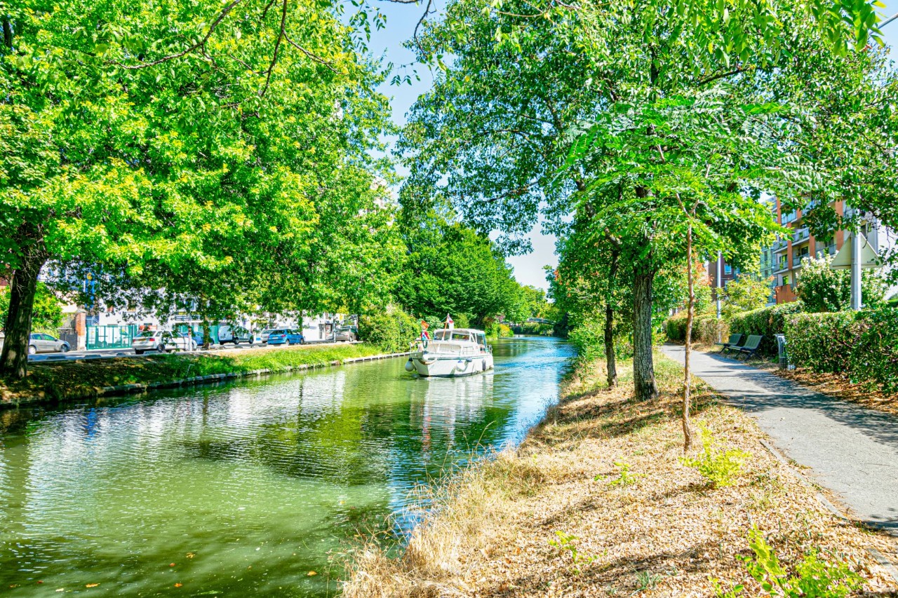 Green, tree-lined canal on which a boat is travelling towards the onlooker. A narrow footpath runs to the right of the canal, with a hedge and buildings to the right.