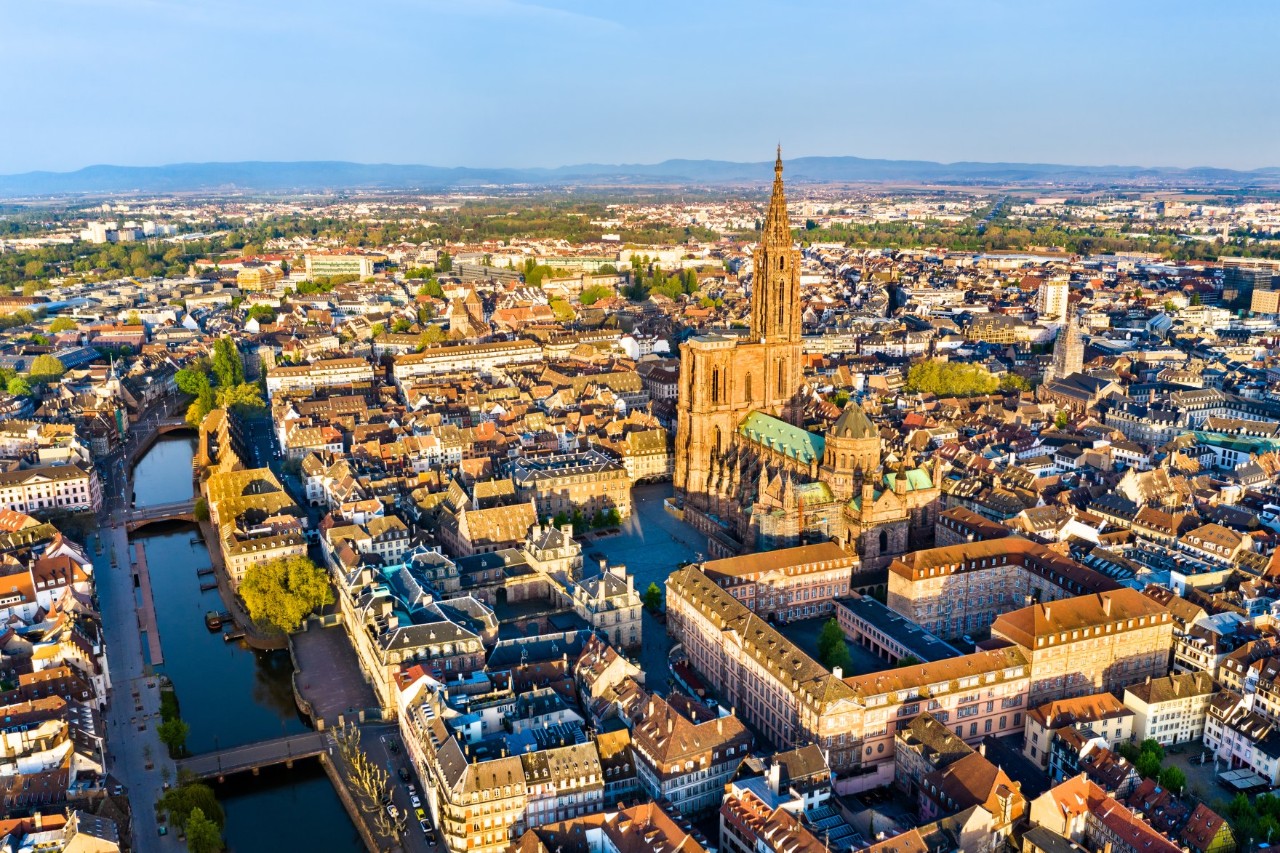 Aerial view with an imposing, tall cathedral in the centre of the picture. Surrounded by the historic buildings of the old town and the branching canals, the city stretches as far as the green hills in the background.
