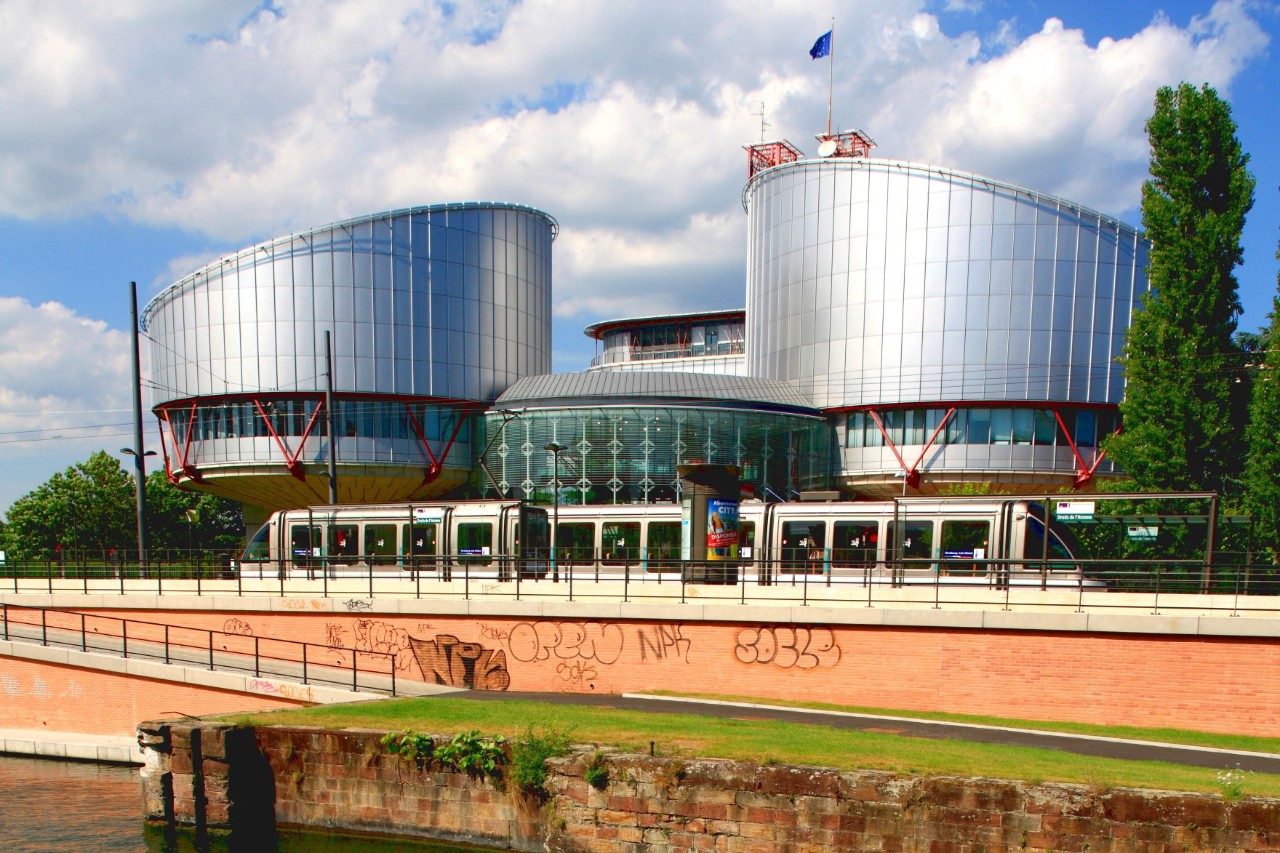 The picture shows a futuristic-looking building complex with round, silver-coloured houses made of glass and metal. In front of it, a tram runs along a wall over a canal. The sky is slightly cloudy.