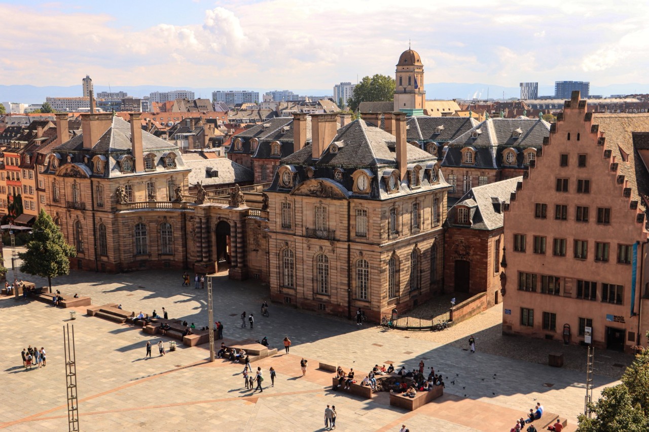 The picture shows a historic square with a large baroque building with ornate decorations, many windows and mansard roofs. The town stretches out in the background.