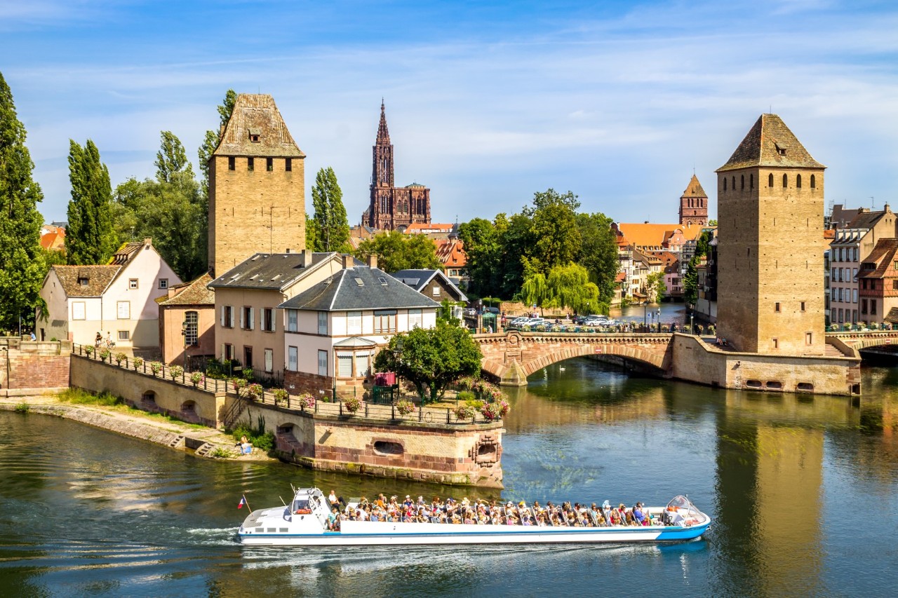 The picture shows a river landscape with a busy excursion boat travelling through calm waters. Behind the boat are two medieval watchtowers and a bridge with two arches crossing the river. In the background, an old town with traditional buildings and the striking Strasbourg Cathedral are visible. The sky is blue.