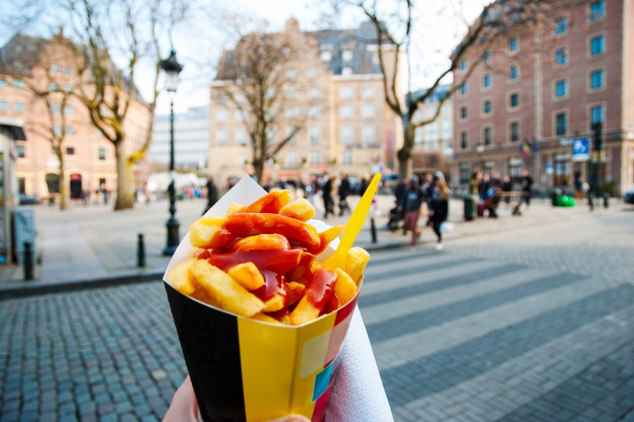 : Belgian fries with ketchup in a bag, busy square in the background, bare trees, brown buildings 