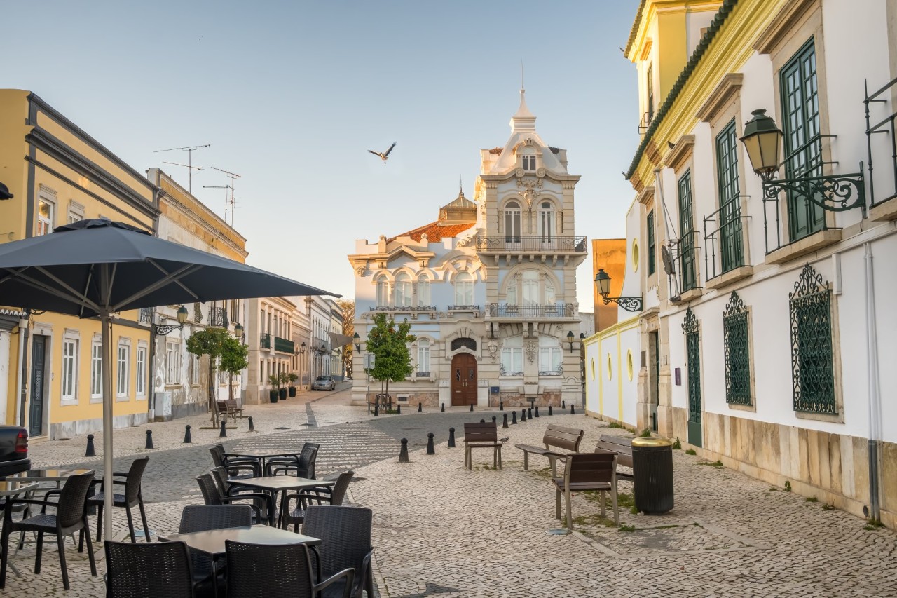 Belmarco mansion in the city center, in front of it a street cafe and benches, parasol, white houses, empty square and flying seagull.  