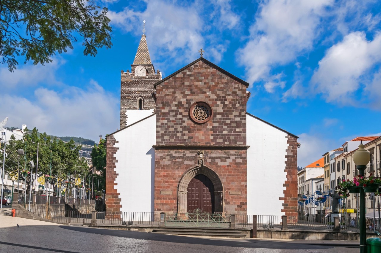White cathedral with brown tower and brick wall, surrounded by buildings and trees 