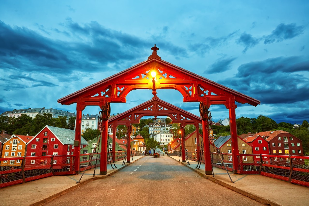 Head-on view of a road leading over a bridge in the evening. Red, high wooden arches cross the bridge road. On the other side of the bridge you can see buildings with colourful facades and the city.   