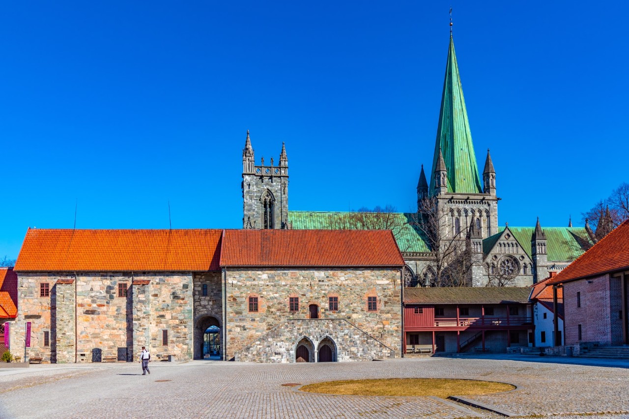 View of a large, empty square bordered by an old stone building with a red roof and a cathedral behind it.  © dudlajzov/stock.adobe.com 