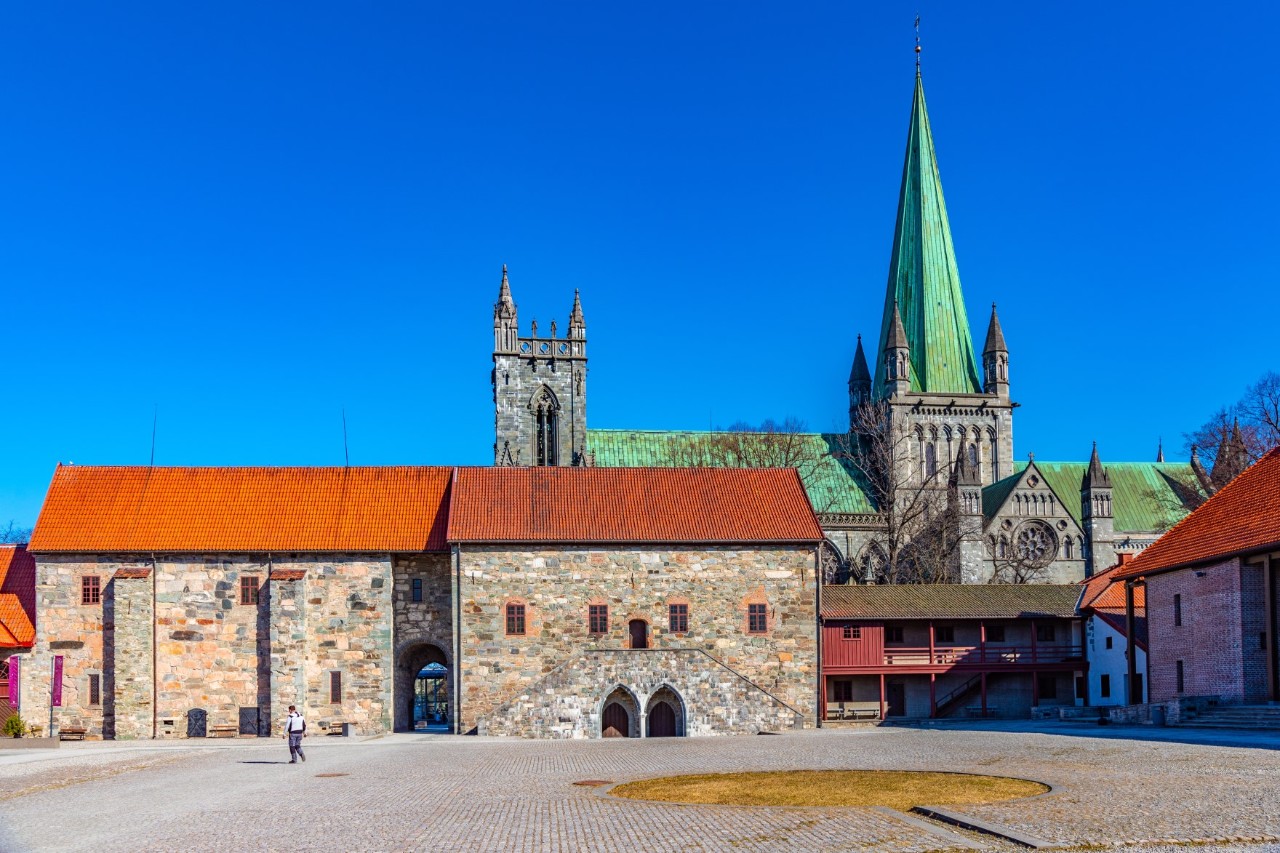 View of a large, empty square bordered by an old stone building with a red roof and a cathedral behind it.  