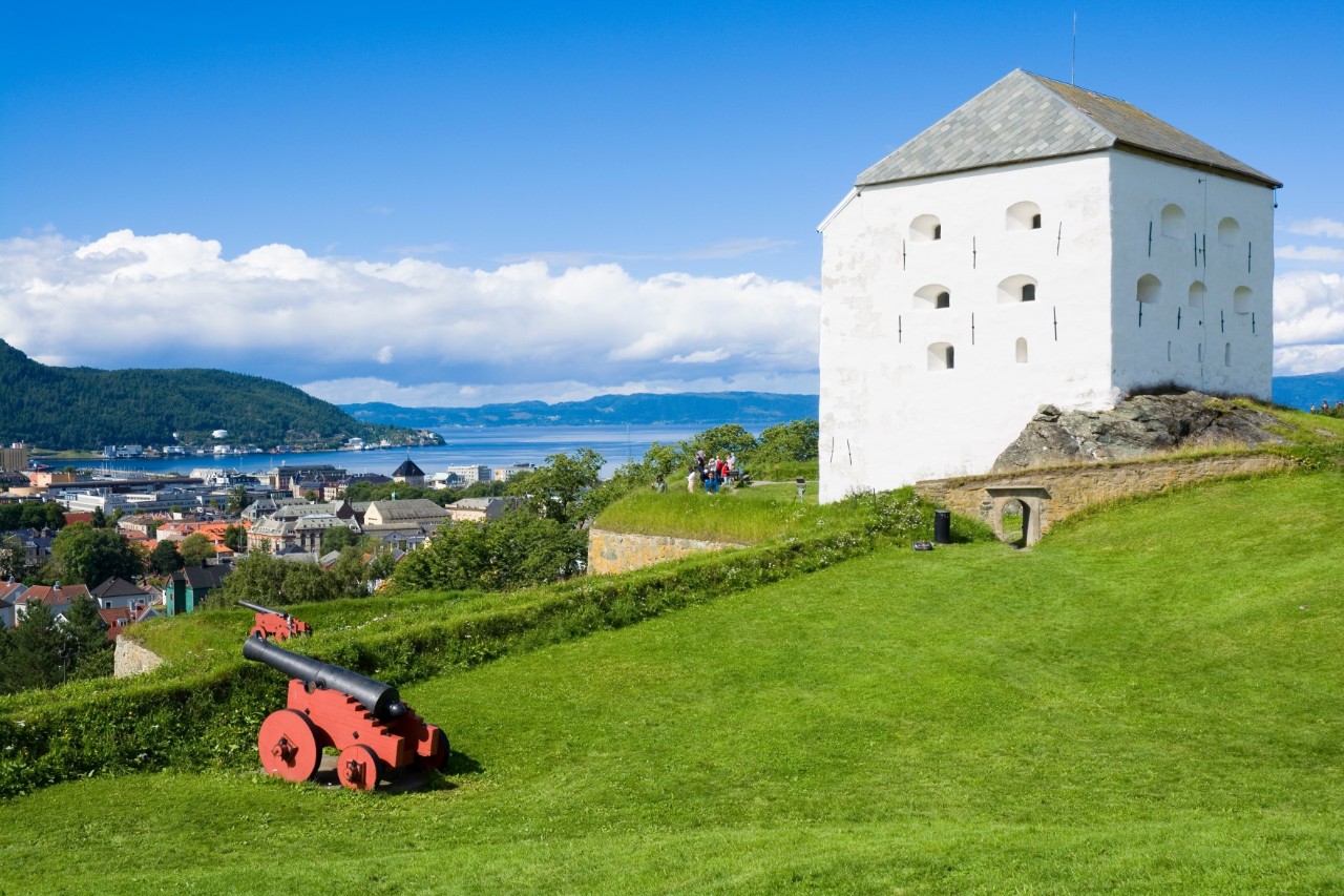White, strong, angular tower building on the right side of the picture, in front of it is a green meadow with an old cannon on the left side of the picture. Buildings, water, mountains in the background.  © Mariusz Switulski/stock.adobe.com 