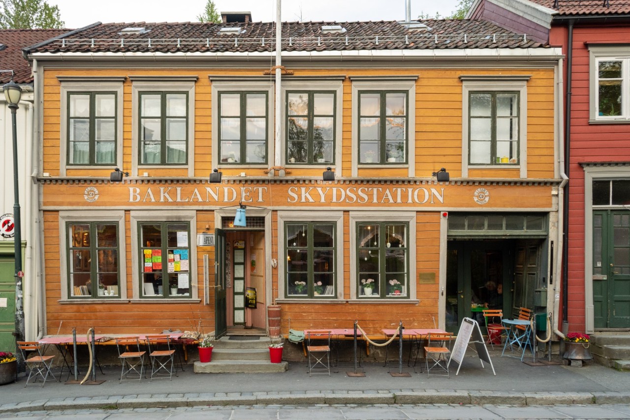 View of a yellow wooden house called “Baklandet Skydsstation” with tables and benches in front of the building. 