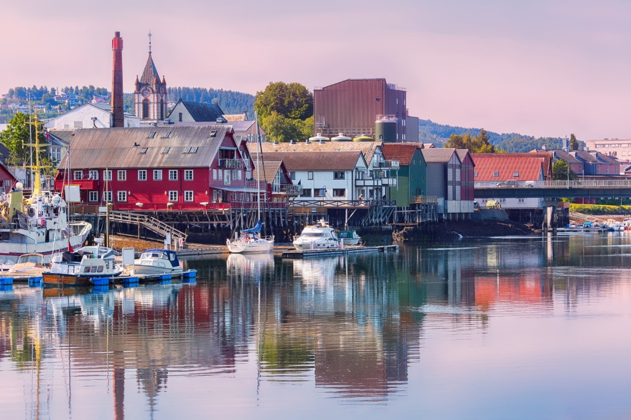 View of a town from the water. Sailing boats are moored on the shore and there are houses with colourful fronts. A church tower and industrial buildings are in the background. 
