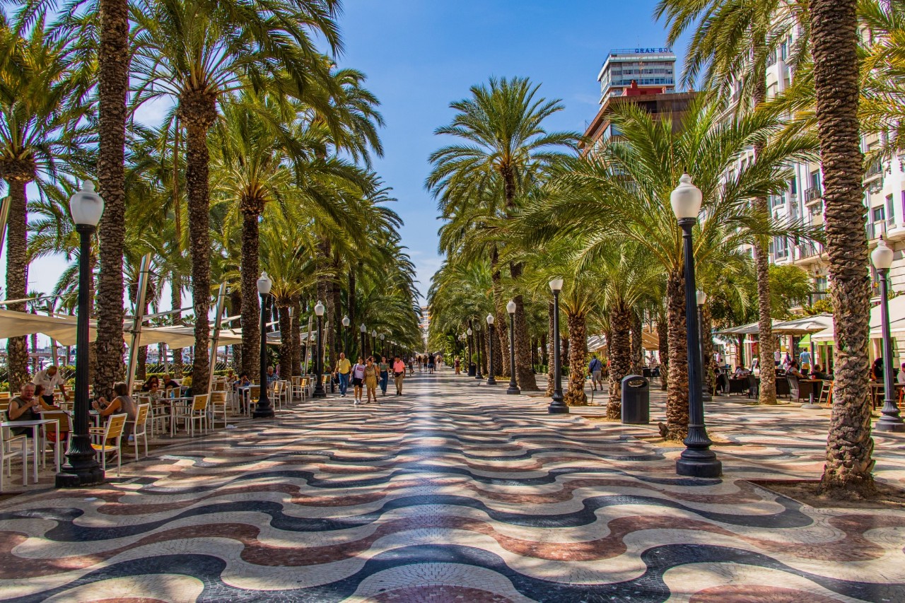 View of a promenade lined with palm trees, the floor of which is made up of a wavy mosaic in the colours ochre, terracotta and black. Black street lamps stand between the palm trees. A street café and a few people strolling can be seen on the left of the picture. A row of buildings can be seen behind the palm trees on the right.